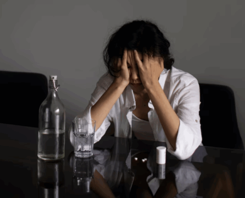 A lesbian person looking confused and anxious sitting on a table with scattered white pills and water.