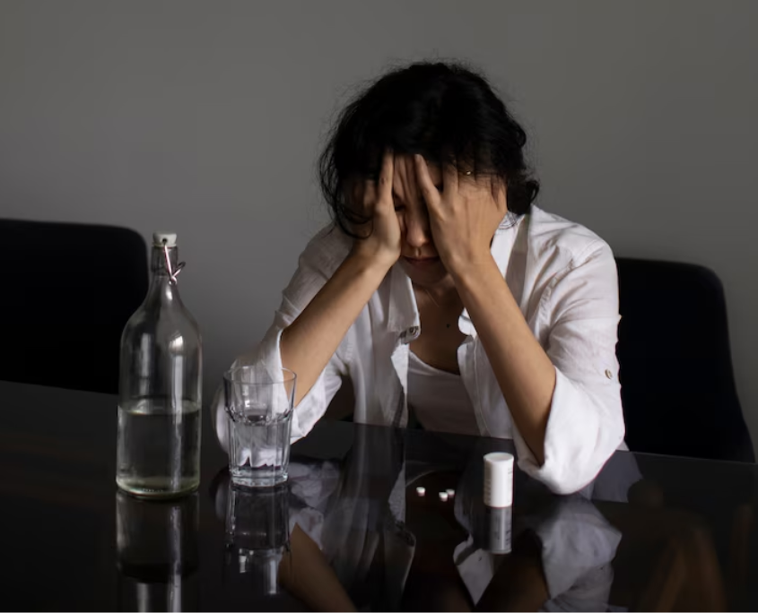 A lesbian person looking confused and anxious sitting on a table with scattered white pills and water.