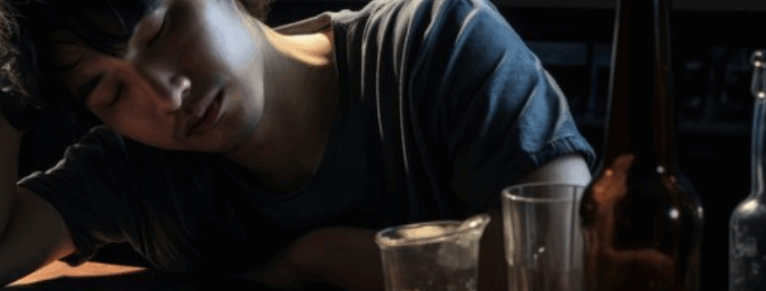 An asian looking young gay man, seemingly drunk in a table with glasses and bottles of alcohol.