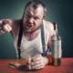 A raging alcoholic yelling while pointing, holding a glass of whiskey next to a bottle.