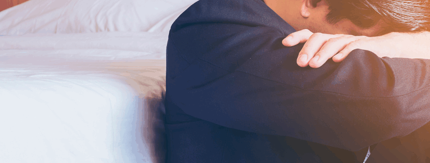 A gay person sitting beside a bed with head down and arms crossed in a type of depression.